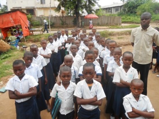 Pupils in white and blue uniforms lined up in rows in the schoolyard with their teachers
