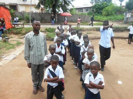 Pupils in white and blue uniforms lined up in rows in the schoolyard with their teachers