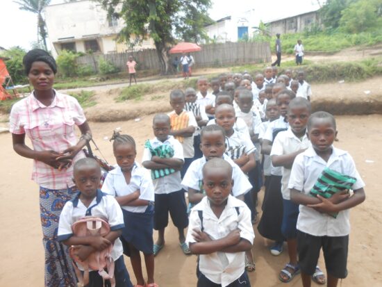 Pupils in white and blue uniforms lined up in rows in the schoolyard with their teachers