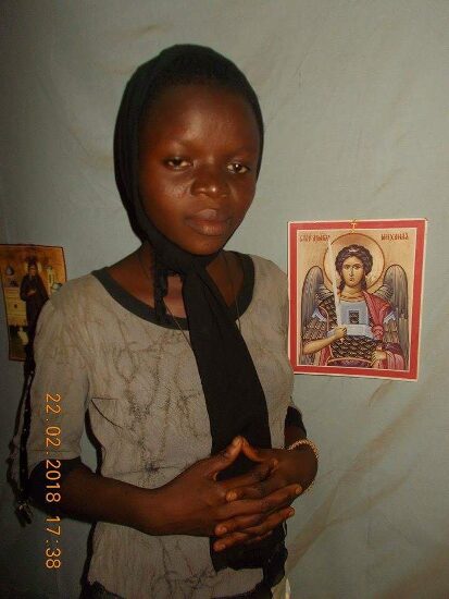 A girl in a head scarf standing before an icon of the Archangel Michael