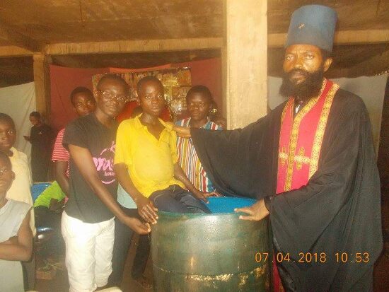 Father Theophile baptizing by immersion in a large barrel font, the faithful looking on