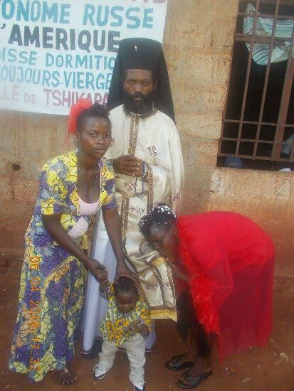 Father Theophile in vestments with newly baptized and their families in front of the church sign of the Parish of the Dormition in Tshikapa