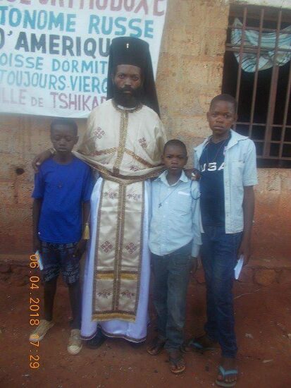 Father Theophile in vestments with newly baptized and their families in front of the church sign of the Parish of the Dormition in Tshikapa