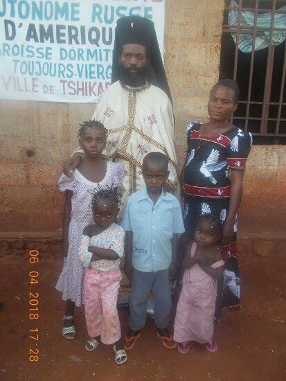 Father Theophile in vestments with newly baptized and their families in front of the church sign of the Parish of the Dormition in Tshikapa