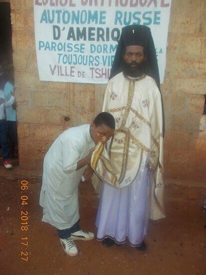 Father Theophile in vestments with newly baptized and their families in front of the church sign of the Parish of the Dormition in Tshikapa