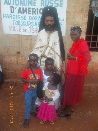 Father Theophile in vestments with newly baptized and their families in front of the church sign of the Parish of the Dormition in Tshikapa