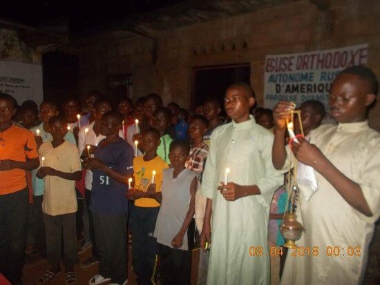 Young men in white robes, one with the censer, among the faithful with candles
